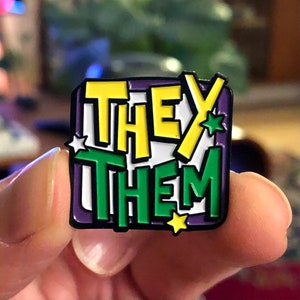 May include: A purple, green, yellow and white enamel pin with the words "THEY THEM" in a bold, blocky font.
