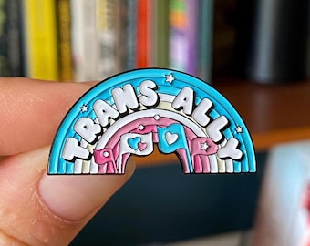 May include: A rainbow-shaped enamel pin with the words "TRANS ALLY" in white. The pin features a light blue outer border, a pink and white rainbow, and small flags with hearts. The pin is held by a person.