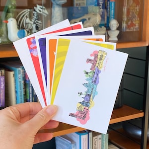 Könnte beinhalten: Eine Hand hält einen Stapel bunter Postkarten. Jede Karte zeigt ein anderes Design, darunter eine Stadtlandschaftsillustration und abstrakte Muster. Die Karten sind weiß mit farbenfrohen Akzenten, das Thema ist Reisen und Kunst.