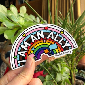 Puede incluir: Una pegatina en forma de arcoíris con las palabras "I AM AN ALLY". El diseño incluye corazones, estrellas y banderas. La firma del artista, "Sophie Green", está en la esquina inferior derecha.