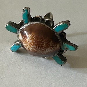 Könnte beinhalten: Silberfarbener Ring in Schildkrötenform mit braun-weißem Muschelmittelteil und türkisfarbenen Akzenten an den Flossen. Der Ring hat ein detailliertes, strukturiertes Design und zeigt eine einzigartige, handgefertigte Ästhetik.