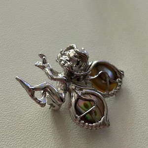 Op de afbeelding: Een zilverkleurige feeën broche met iriserende vleugels. De fee zit met uitgestrekte armen en benen. De vleugels zijn een mix van bruin, geel en groen. De broche is waarschijnlijk van metaal.