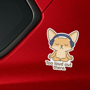 May include: A cartoon fox wearing headphones sits in a meditative pose with the text "Too loud out there." printed below. The fox is light brown with a white belly and blue headphones. The background is a red car.