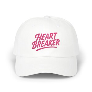 May include: A white baseball cap with the embroidered text "HEART BREAKER" in a cursive, pink font. The cap has a curved brim and a rounded crown, suitable for casual wear.