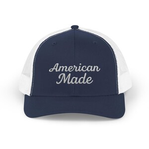 Puede incluir: Gorra de camionero azul marino y blanca con el texto bordado "American Made". La gorra tiene una visera curva y una parte trasera de malla. Un accesorio de moda para el uso diario.