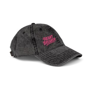 May include: A distressed black baseball cap with the text "HEART BREAKER" embroidered in pink. The cap has a curved brim, a button on top, and an adjustable strap at the back. The hat is made of a soft, worn material.