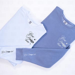 May include: Two long-sleeved sweatshirts, one light blue and one periwinkle, are displayed on a white surface. Each shirt has the text "Your Design here" printed on the front and sleeve. The shirts are folded neatly, showcasing the design.