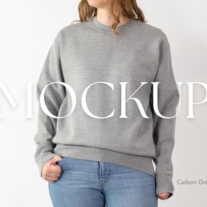 以下が含まれることがあります： 前面に大きな白い文字で「MOCKUP」という文字が入ったグレーのクルーネックのトレーナー。トレーナーはカーボングレー色で、ブルージーンズと合わせています。「Carbon Grey」の文字が右下にあります。