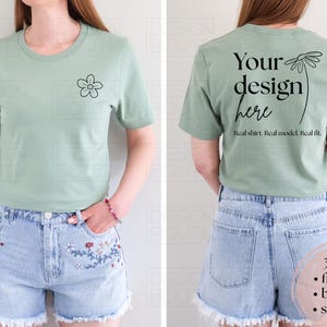 Op de afbeelding: Saliegroen t-shirt met een kleine bloem op de voorkant. De achterkant van het shirt heeft de tekst "Your design here" met een madeliefjesillustratie. Het model draagt een spijkerbroekshort.