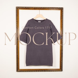 May include: A charcoal grey short-sleeve t-shirt hanging on a light-coloured hanger inside a decorative gold-toned frame. The text Comfort Colors 1717 and the word MOCKUP are overlaid on the image in a light beige colour.