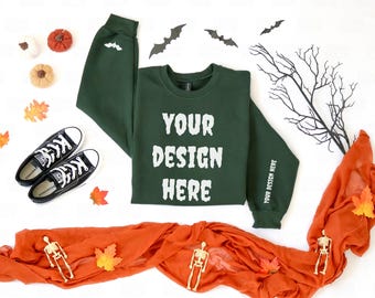 May include: A dark green sweatshirt with the text "YOUR DESIGN HERE" in white. The sleeves have a bat graphic and the text "YOUR DESIGN HERE". Black sneakers, decorative pumpkins, and fall leaves are also present, along with a decorative orange scarf and skeletons.
