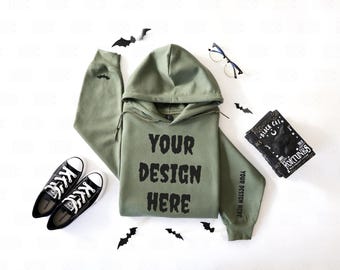 May include: An olive green hoodie with the text "YOUR DESIGN HERE" in black, black Converse sneakers, a book with the text "BLACK CAT FORTUNES", and black bat decorations. A pair of glasses is also present.
