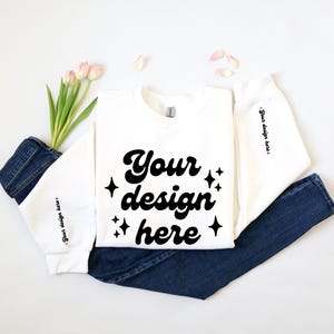 Könnte beinhalten: Weißer Langarm-Sweatshirt mit dem Text "Your design here" in schwarzer Retro-Schrift mit Sternen. Der Sweatshirt ist gefaltet und auf einer weißen Oberfläche mit einer blauen Jeans und rosa Blumen gelegt.
