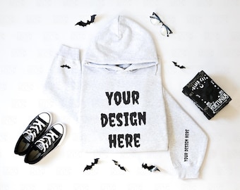 May include: A light gray hoodie with the text "YOUR DESIGN HERE" in black. Black Converse sneakers, a black bandana with text, and bat-shaped decorations are arranged around the hoodie. A pair of glasses is also present.