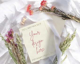Può includere: Un mockup di biglietto 5x7 con il testo "Your design here" in un carattere corsivo. Il biglietto è circondato da fiori secchi e una bobina di legno su uno sfondo di tessuto bianco. L'immagine è una posa piatta.