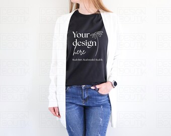 May include: A black t-shirt with the text "Your design here" in white, paired with blue jeans and a white cardigan. The shirt also has the text "Real shirt. Real model. Real fit."