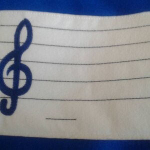May include: White fabric patch with a blue treble clef embroidered on the left side. The patch has black lines sewn across it, resembling a music staff.