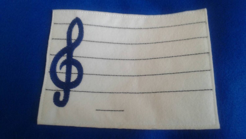 May include: White fabric patch with a blue treble clef embroidered on the left side. The patch has black lines sewn across it, resembling a music staff.