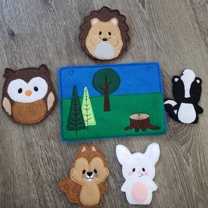 May include: A felt play set with a blue background and green grass. The set includes a brown owl, a brown squirrel, a white rabbit, a black and white skunk, and a brown hedgehog. The set also includes a green tree and a brown tree stump.