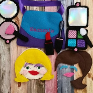 May include: A felt play set for pretend play, featuring a purple purse with the name "Sammie" embroidered on it, a pink compact mirror, a red lipstick, a black mascara wand, a blue eyeshadow palette, and two felt faces with blonde and brown hair.