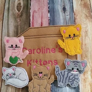 May include: Five felt cat appliques with embroidered details. The appliques are in various colors, including pink, yellow, gray, and brown. The appliques are arranged on a brown felt background with the text "Caroline's Kittens" embroidered in pink thread.