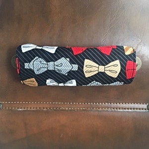 May include: A rectangular eyeglasses case with a black background and white pinstripes. The case is decorated with various bow tie designs in red, white, gray, and tan. The case is placed on a brown leather surface.
