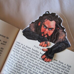 Puede incluir: Un marcap&aacute;ginas con una ilustraci&oacute;n detallada de un personaje con pelo largo y oscuro, barba y ojos azules, mirando por encima de las p&aacute;ginas de un libro. El personaje lleva una armadura y tiene una expresi&oacute;n severa. El marcap&aacute;ginas es de pl&aacute;stico transparente.