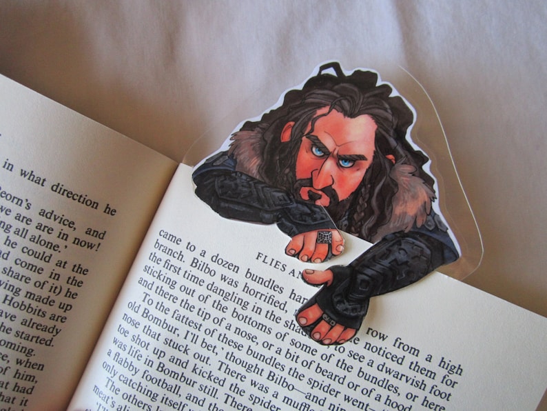 Puede incluir: Un marcap&aacute;ginas con una ilustraci&oacute;n detallada de un personaje con pelo largo y oscuro, barba y ojos azules, mirando por encima de las p&aacute;ginas de un libro. El personaje lleva una armadura y tiene una expresi&oacute;n severa. El marcap&aacute;ginas es de pl&aacute;stico transparente.