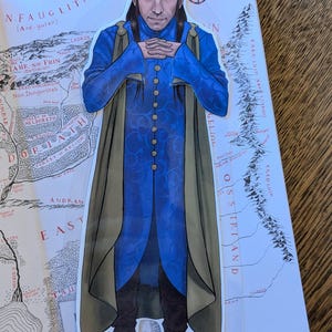 Può includere: Un segnalibro con un personaggio in un abito blu e verde oliva, con una corona d'oro. Il segnalibro è posto su una mappa con testo tra cui "NFAUGLITH" e "EAST".