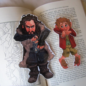 Puede incluir: Dos recortes de personajes de una novela de fantasía, colocados en un libro abierto. Un personaje es un enano con una larga barba y un abrigo forrado de piel. El otro es un hobbit con una chaqueta roja y pantalones marrones.