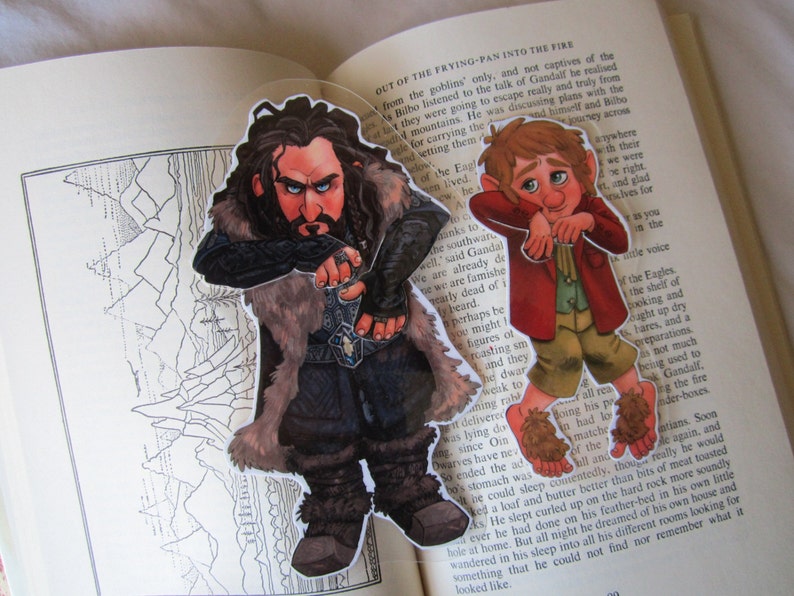 Puede incluir: Dos recortes de personajes de una novela de fantas&iacute;a, colocados en un libro abierto. Un personaje es un enano con una larga barba y un abrigo forrado de piel. El otro es un hobbit con una chaqueta roja y pantalones marrones.