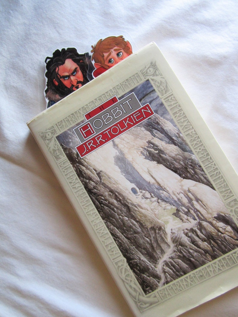 Puede incluir: Un ejemplar de "El Hobbit" de J.R.R. Tolkien con un marcap&aacute;ginas con ilustraciones de dos personajes. El libro tiene una ilustraci&oacute;n de paisaje en la portada.