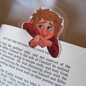 Puede incluir: Un marcap&aacute;ginas transparente con una ilustraci&oacute;n de dibujos animados de un personaje con pelo rojo y una chaqueta roja, colocado en un libro. El personaje tiene una expresi&oacute;n pensativa. El marcap&aacute;ginas est&aacute; en una p&aacute;gina con texto impreso.