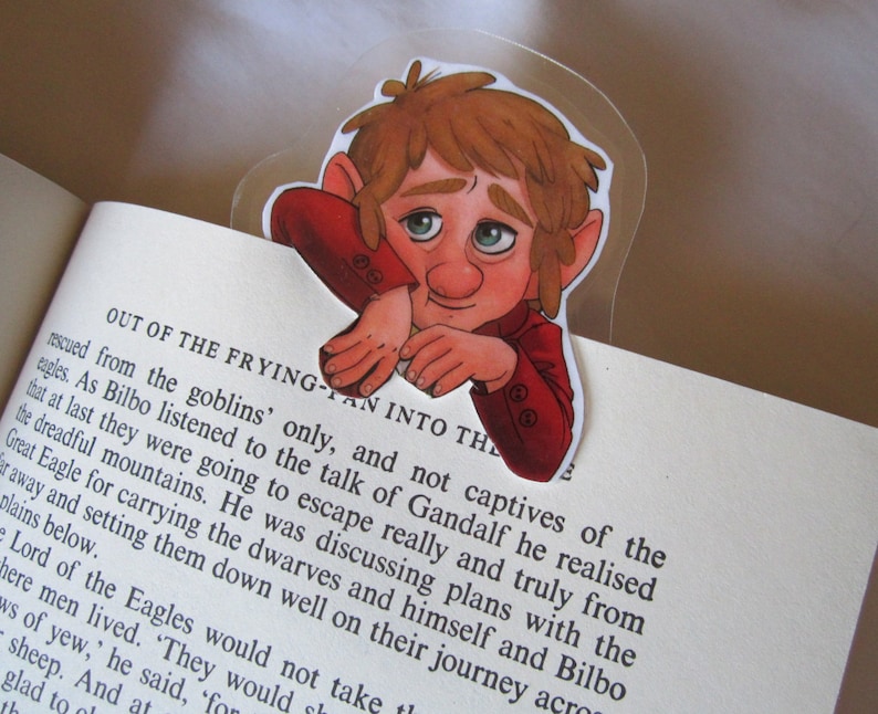 Puede incluir: Un marcap&aacute;ginas transparente con una ilustraci&oacute;n de dibujos animados de un personaje con pelo rojo y una chaqueta roja, colocado en un libro. El personaje tiene una expresi&oacute;n pensativa. El marcap&aacute;ginas est&aacute; en una p&aacute;gina con texto impreso.