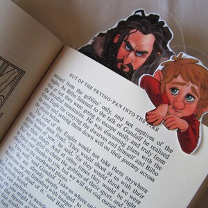 Puede incluir: Dos marcap&aacute;ginas de personajes con ilustraciones de personajes de una novela de fantas&iacute;a. Un marcap&aacute;ginas representa a un hombre con el rostro severo y barba, y el otro a un personaje vestido de rojo con expresi&oacute;n preocupada. Los marcap&aacute;ginas est&aacute;n en un libro abierto.