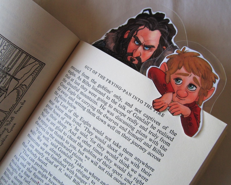 Puede incluir: Dos marcap&aacute;ginas de personajes con ilustraciones de personajes de una novela de fantas&iacute;a. Un marcap&aacute;ginas representa a un hombre con el rostro severo y barba, y el otro a un personaje vestido de rojo con expresi&oacute;n preocupada. Los marcap&aacute;ginas est&aacute;n en un libro abierto.