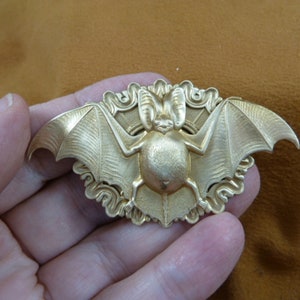 May include: A gold-toned metal bat brooch with intricate details. The bat is depicted with outstretched wings and a detailed body. The brooch is set against a background of swirling, decorative elements.