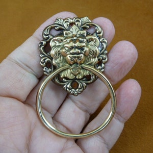 May include: A gold-toned metal ring pull with a lion's head design. The ring pull has a decorative, ornate frame with a detailed lion's face in the center.