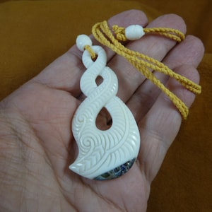 May include: A white bone pendant with intricate carvings in the shape of a stylised fish. The pendant is on a yellow cord and is being held in a hand.