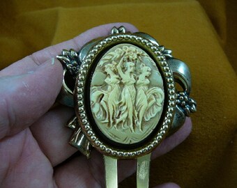 May include: A gold-tone brooch featuring a cameo with three women in relief. The cameo is set in a round frame with a beaded edge. The brooch has a decorative bow design on the back.
