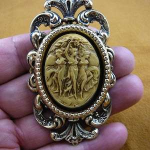 Puede incluir: Un broche de tono dorado con un camafeo ovalado que presenta tres mujeres en un tallado en relieve. El camafeo está rodeado por un borde de cuentas y el broche tiene un diseño decorativo y ornamentado.