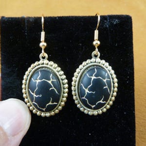 Puede incluir: Un par de pendientes de tono dorado con piedras ovaladas negras. Las piedras tienen un diseño agrietado o veteado.