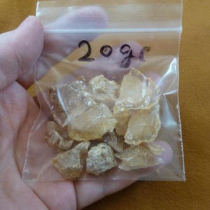 May include: A clear plastic resealable bag containing multiple small, translucent, pale yellow, irregular rock fragments. The bag has the text 20g written in black marker on the front surface, indicating the weight of the contents inside.
