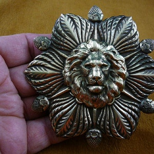 May include: A silver-toned metal lion head brooch with a detailed design of leaves and berries surrounding the head.