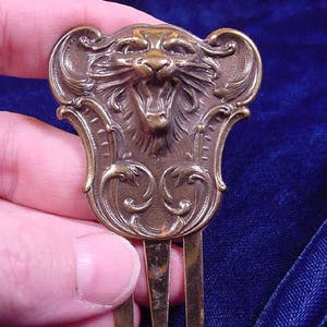Puede incluir: Un peine para el cabello de bronce con un diseño detallado de una cabeza de gato gruñendo con adornos de desplazamiento.