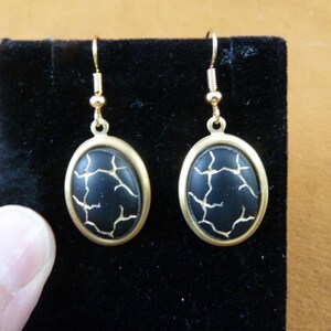 Puede incluir: Un par de pendientes de tono dorado con piedras ovaladas negras. Las piedras tienen un diseño craquelado.