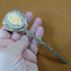May include: A gold-tone hair pick with a heart-shaped head featuring a cameo of a floral design. The handle is decorated with a floral pattern.
