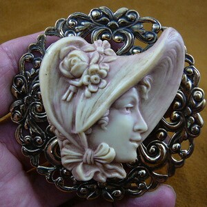 May include: A vintage-style cameo brooch featuring a woman's face in profile, wearing a hat with floral accents. The cameo is set in a gold-toned metal frame with an intricate filigree design.