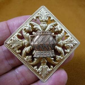 May include: A gold-toned square brooch with an ornate design featuring a stylized vase in the center. The vase is made of copper and has a geometric pattern.