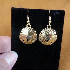 May include: Gold-tone dangle earrings featuring a sand dollar design. The earrings are on a black velvet background.
